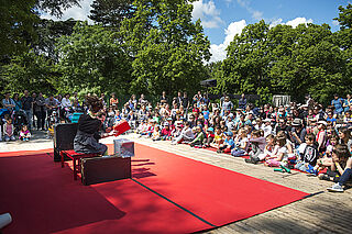 De nombreux spectateurs assistents, assis, à un spectacle joué par deux comédiens sur un grand tapis rouge.